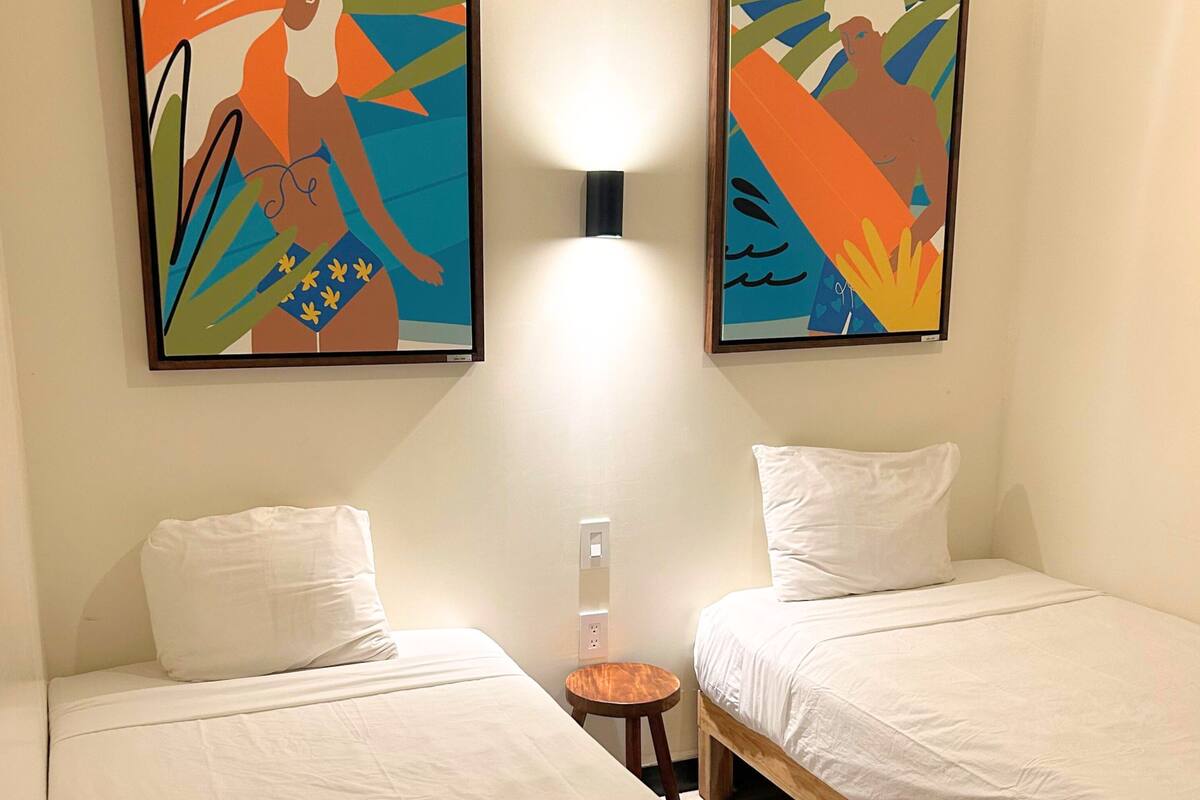 Une chambre d’hôtel avec deux lits simples, une petite table ronde et deux tableaux abstraits aux couleurs vives accrochés au mur.