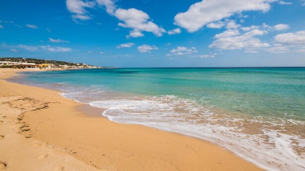 Una spiaggia con acque cristalline turchesi, una costa sabbiosa e alcuni edifici sullo sfondo.