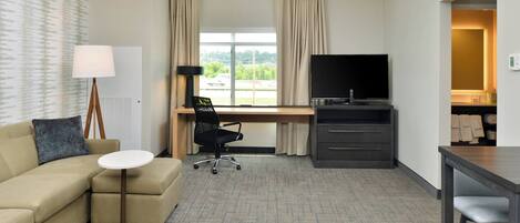 A hotel room with a sofa, a small round table, a desk with a chair, a TV, and a view of the outdoors.