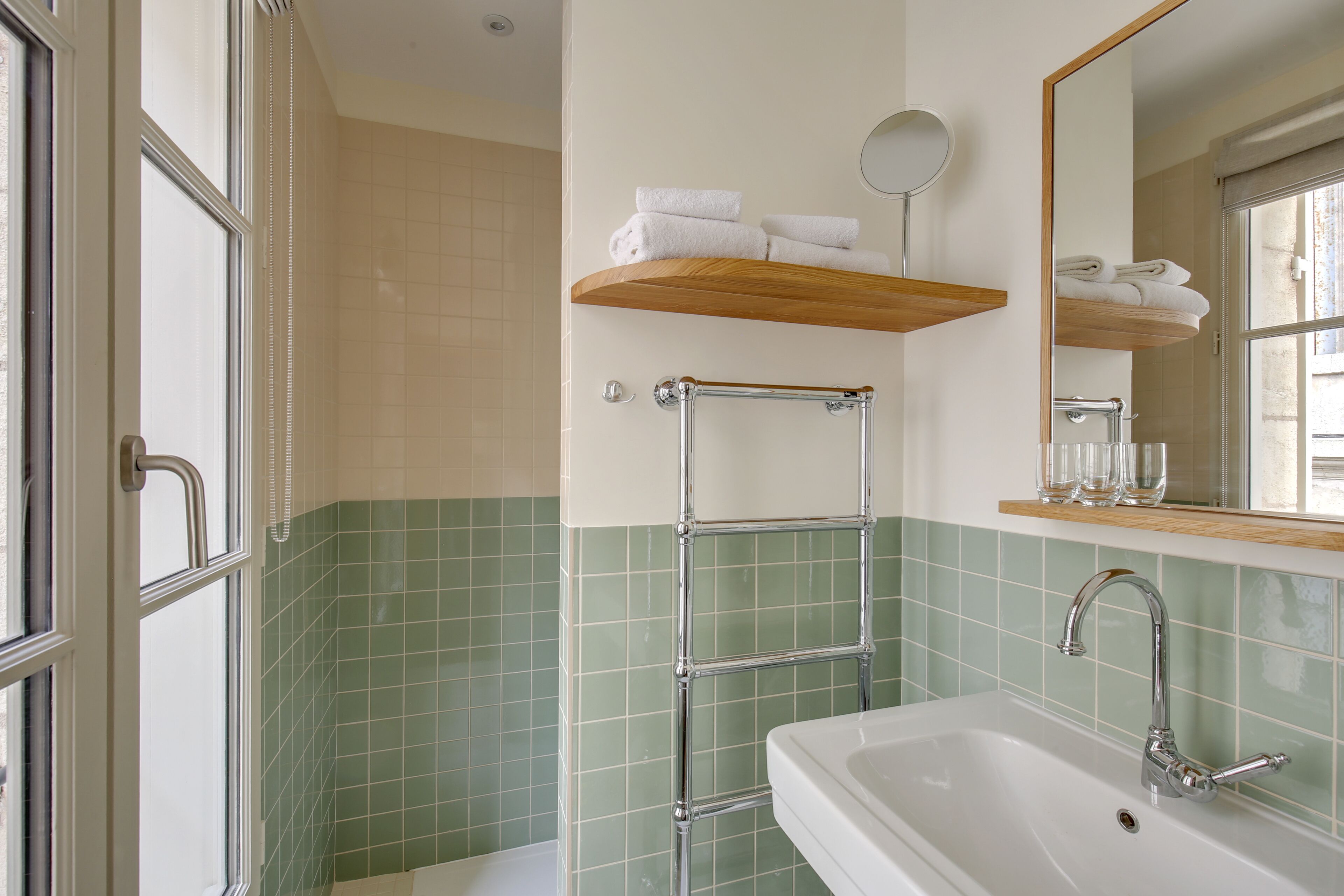 Ein Badezimmer mit einem weißen Waschbecken, grünen Fliesen, einem Holzboden mit Handtüchern, einer Duschkabine aus Glas und einem Spiegel.