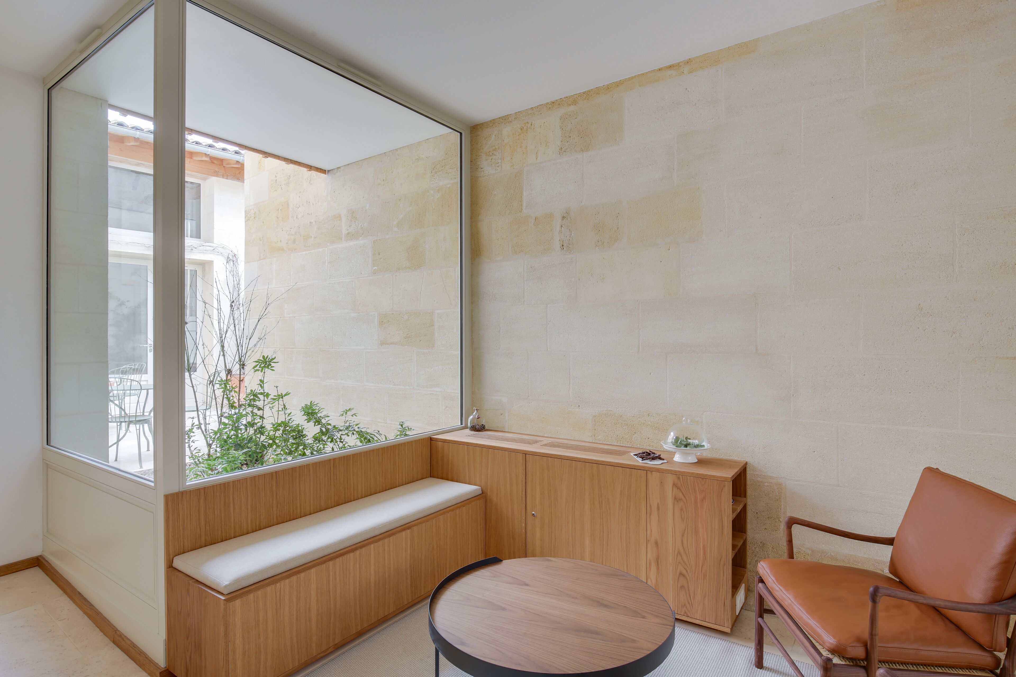Una habitación moderna con un ventanal grande, un banco de madera, una mesa redonda y un sillón de cuero.