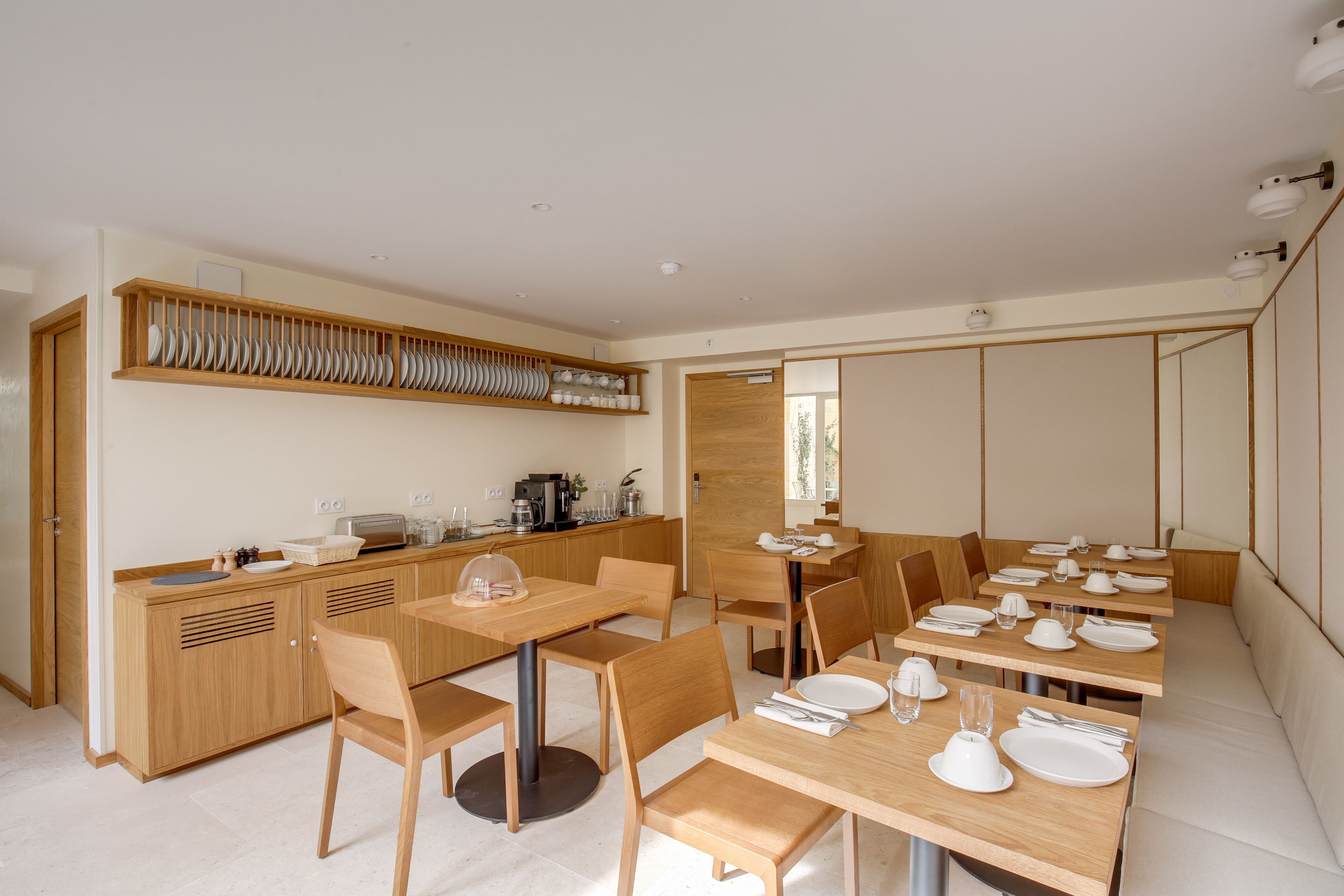 A modern dining area with wooden tables and chairs, a kitchen with a dish rack, and a seating area with cushioned benches.