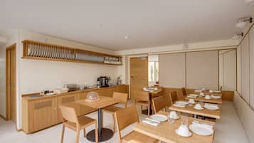 A modern dining area with wooden tables and chairs, a kitchen with a dish rack, and a seating area with cushioned benches.