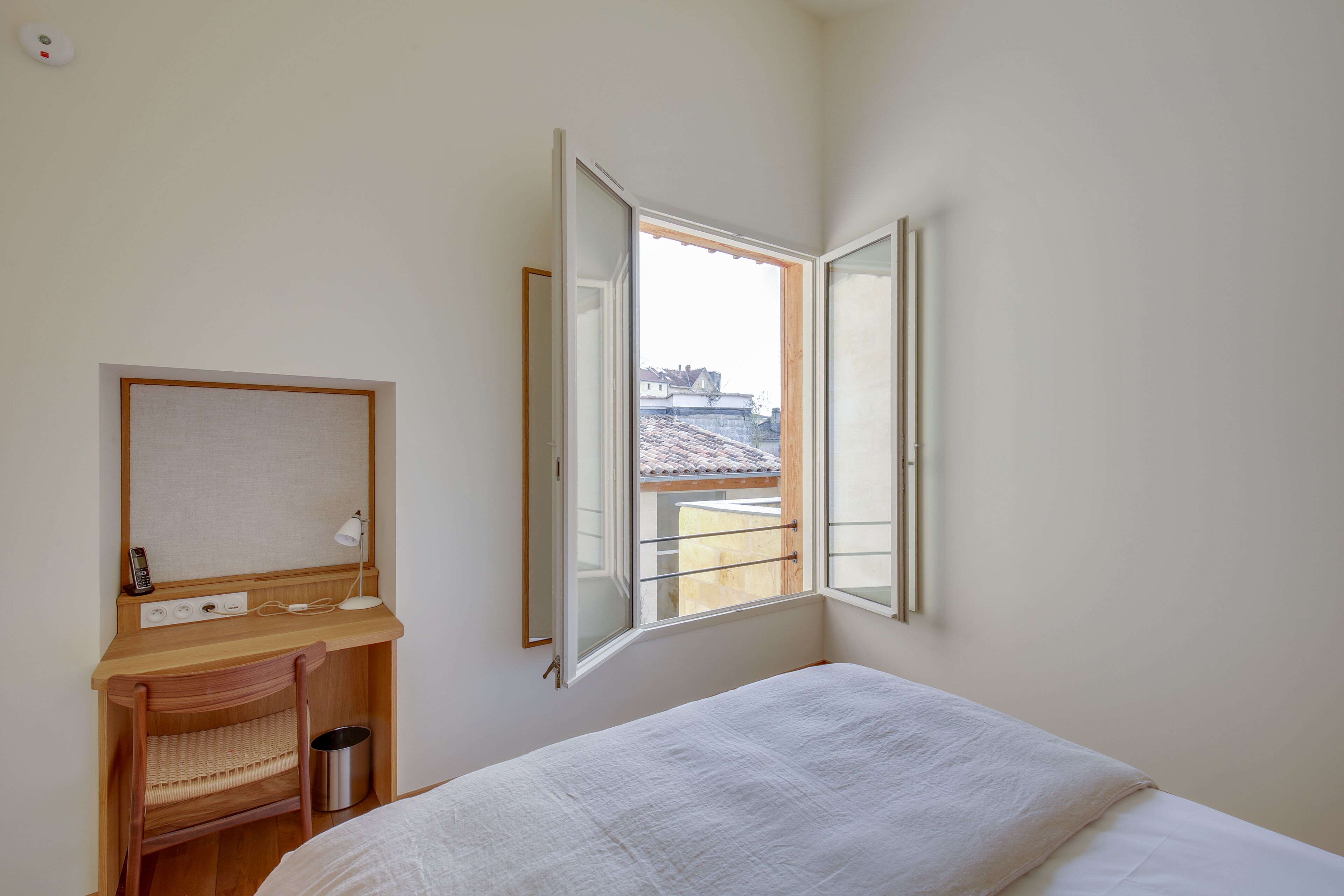 A bedroom with a wooden desk, a chair, a bed, and a window with an open view of buildings.