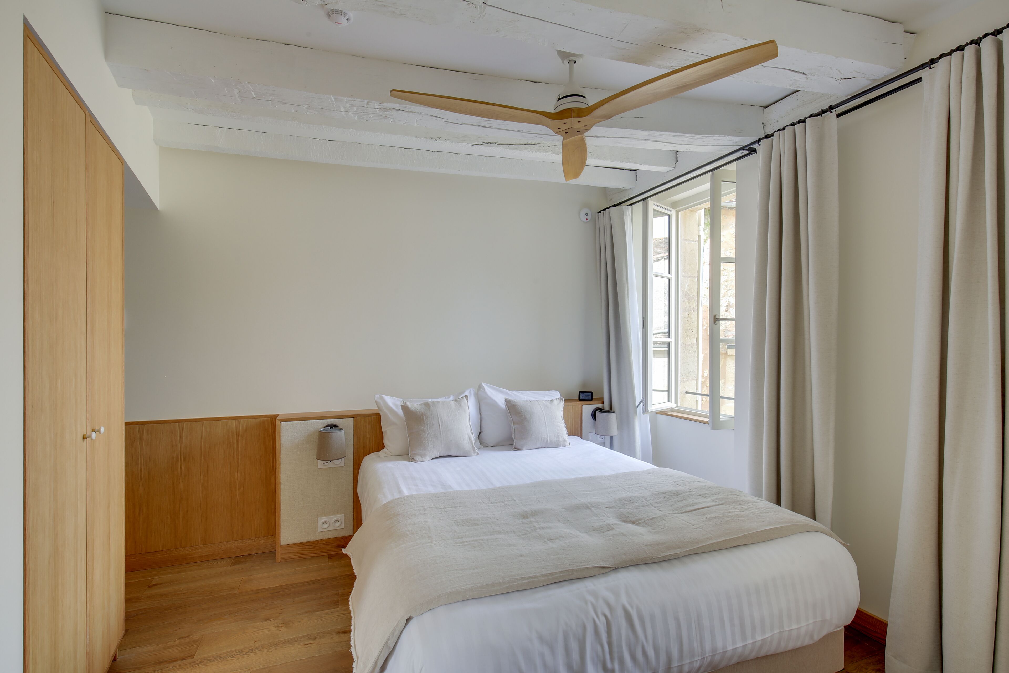 Un dormitorio con un armario de madera, una cama con ropa de cama blanca, un ventilador de techo y una ventana con cortinas.