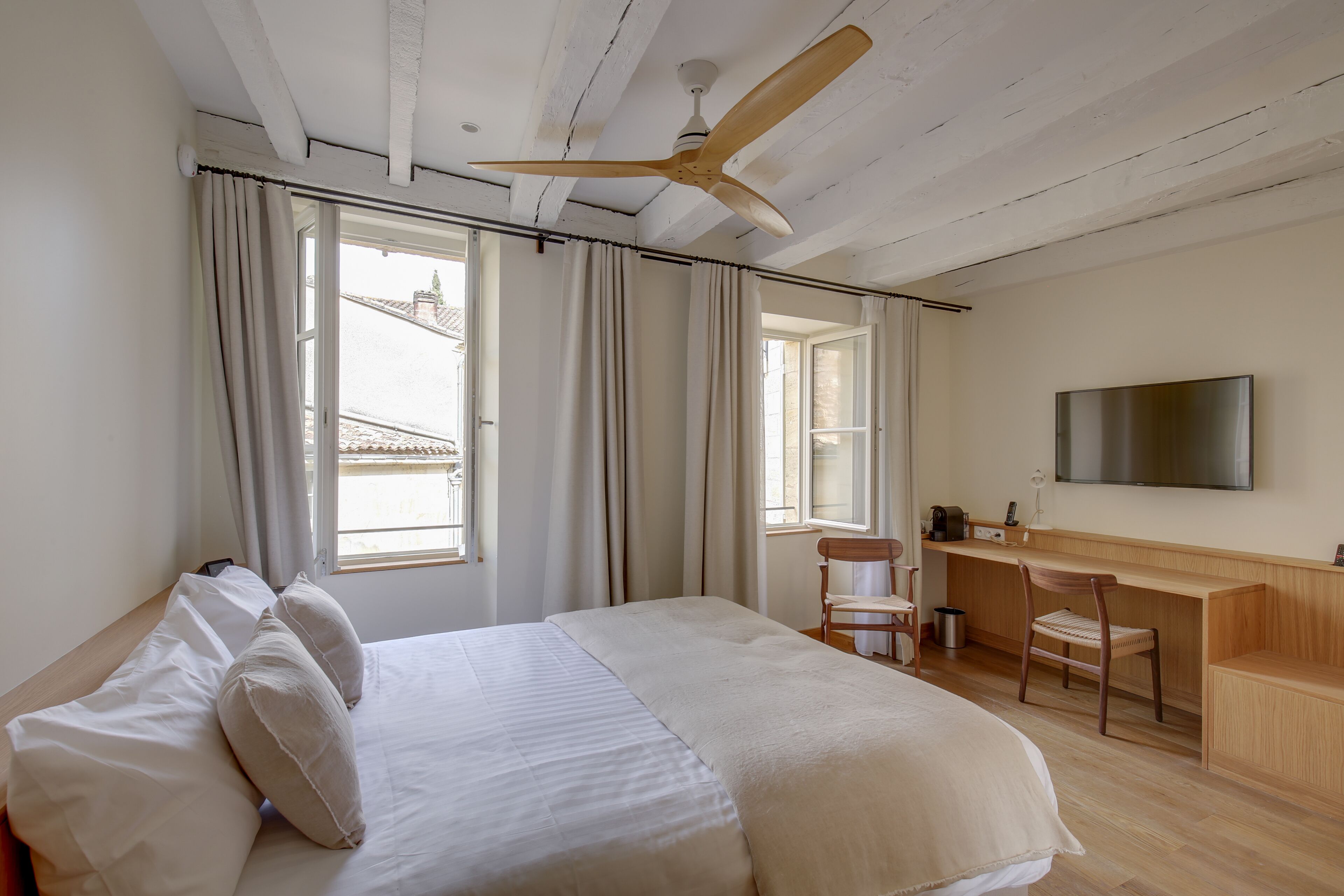 A bedroom with a bed, a desk, a chair, a ceiling fan, and a TV mounted on the wall.
