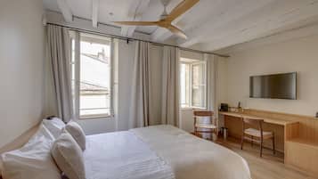 A bedroom with a bed, a desk, a chair, a ceiling fan, and a TV mounted on the wall.