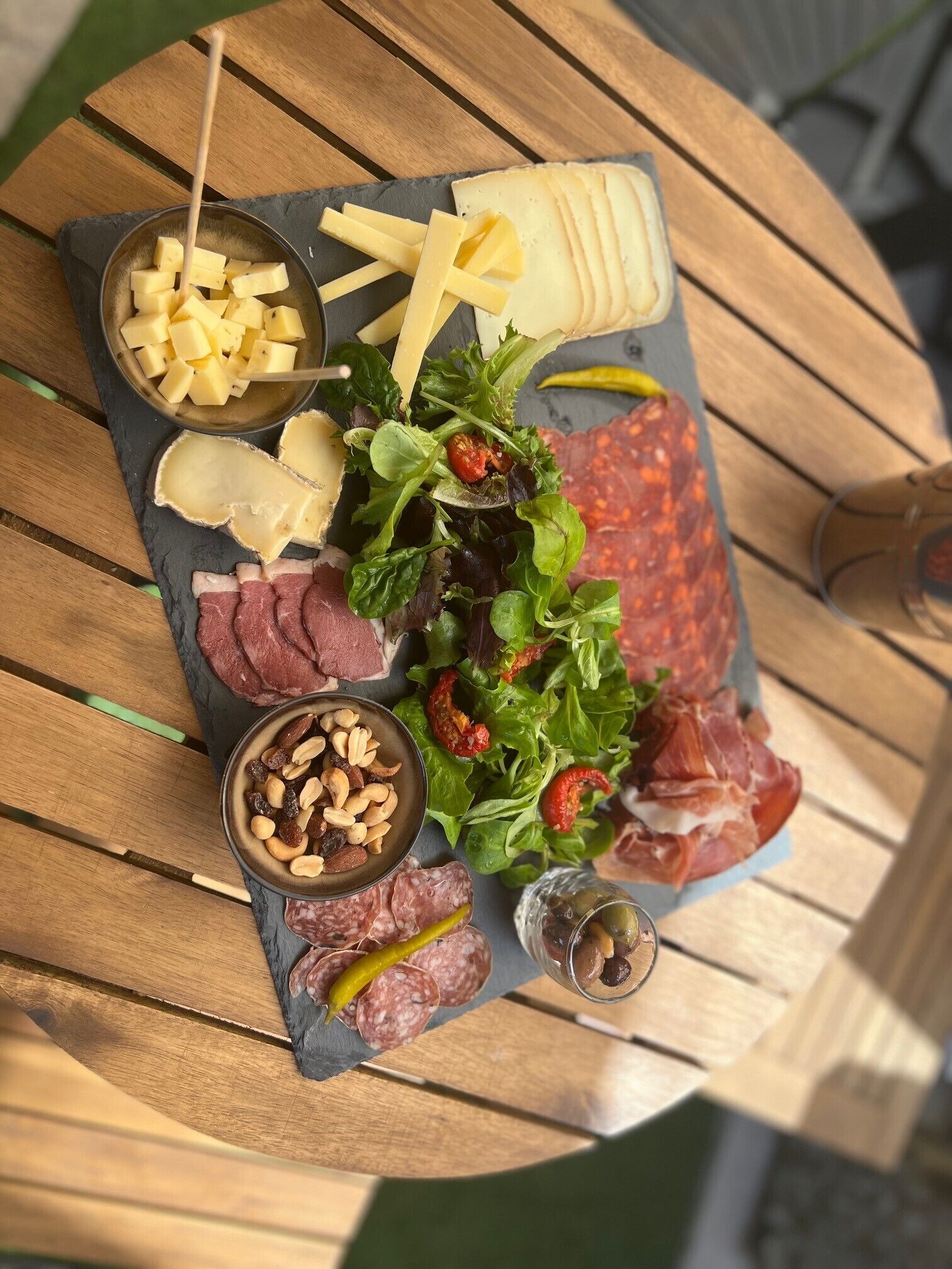 A charcuterie board with various meats, cheeses, nuts, and pickled vegetables on a slate board.