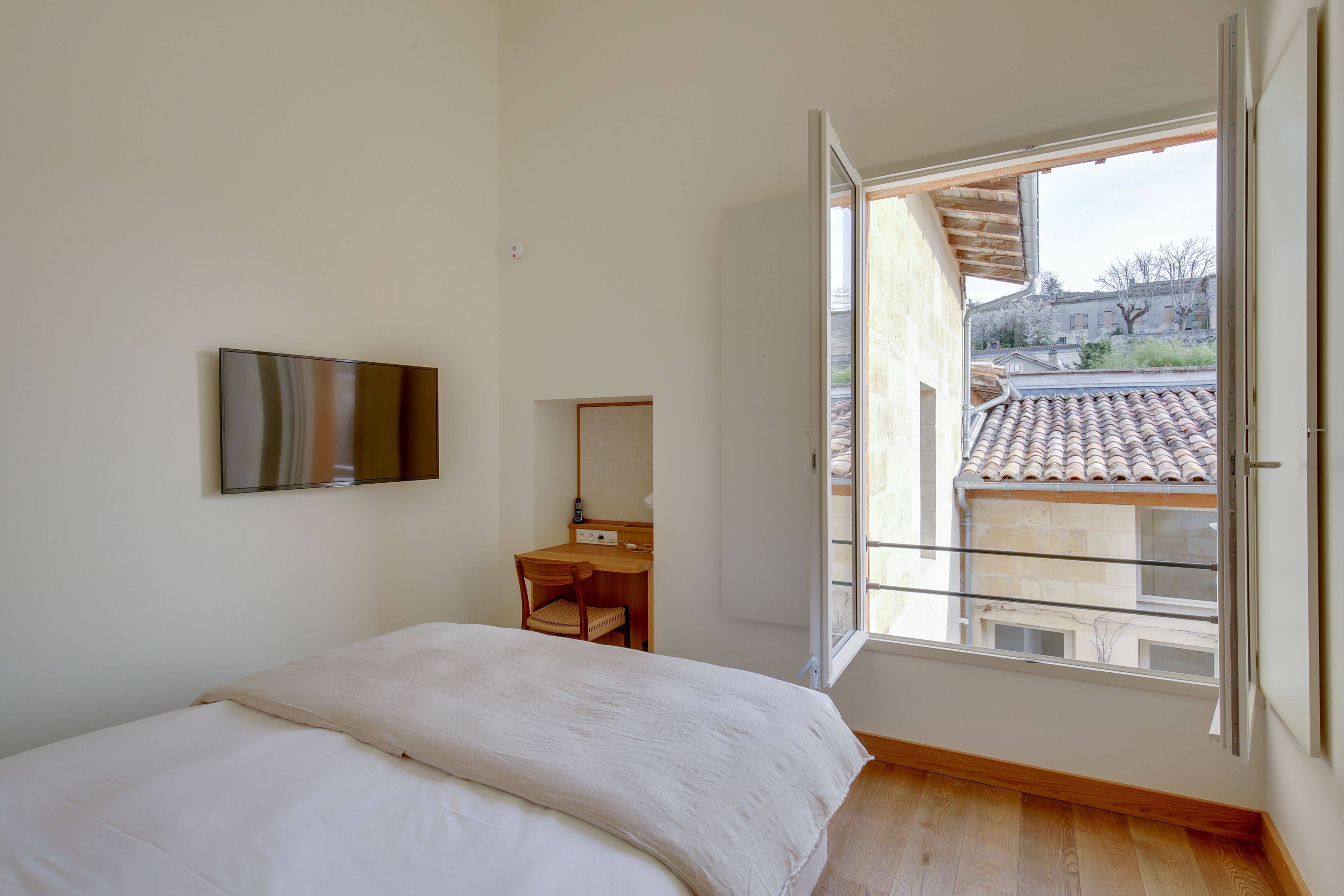 A bedroom with a bed, a desk, a chair, and a TV on the wall. There is a window with a view of a building outside.