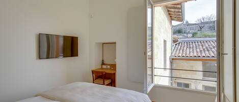 Un dormitorio con cama, escritorio, silla y televisor en la pared. Se ve un edificio a través de la ventana.