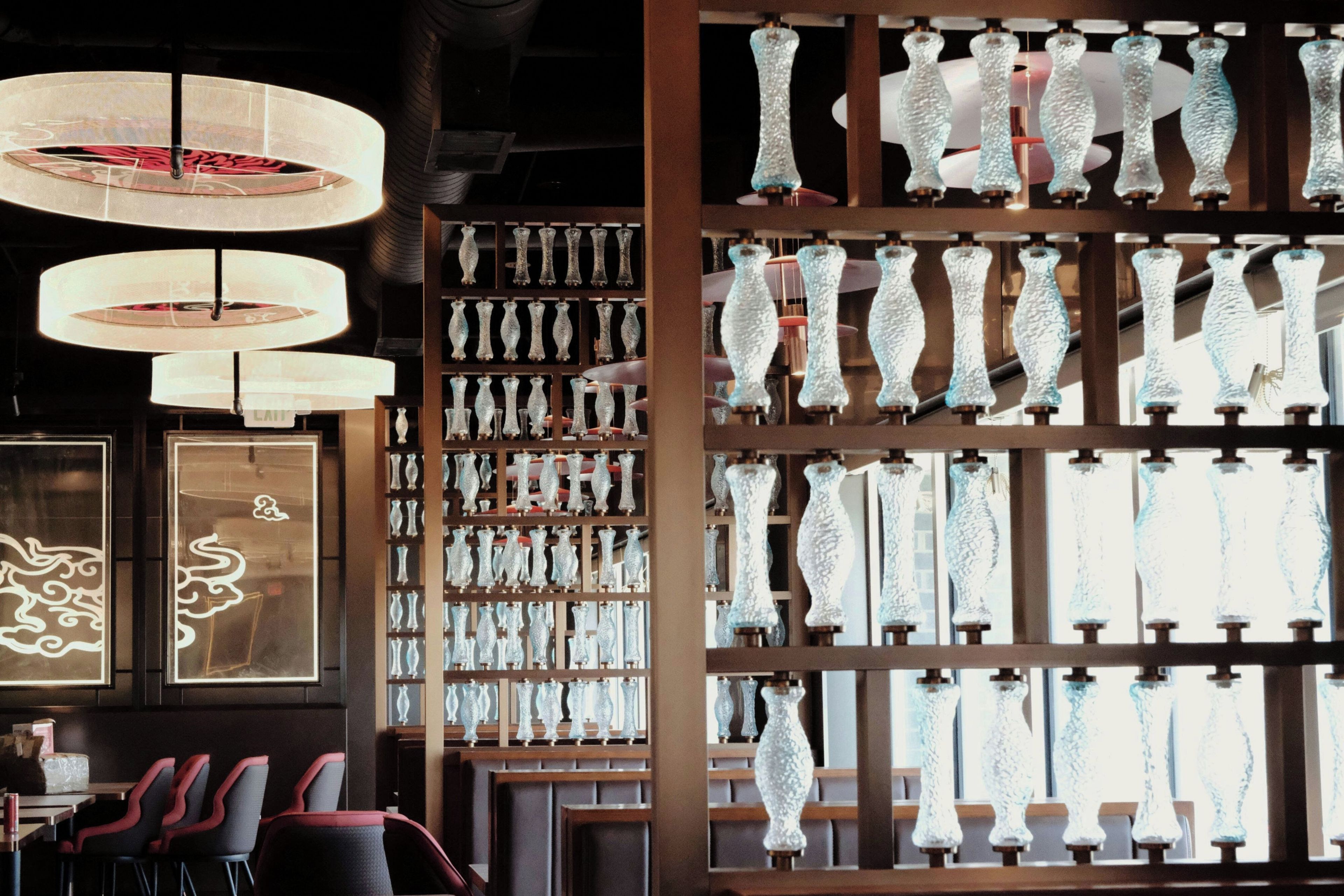 A restaurant interior with hanging pendant lights, a wall of glassware, and a seating area with chairs.