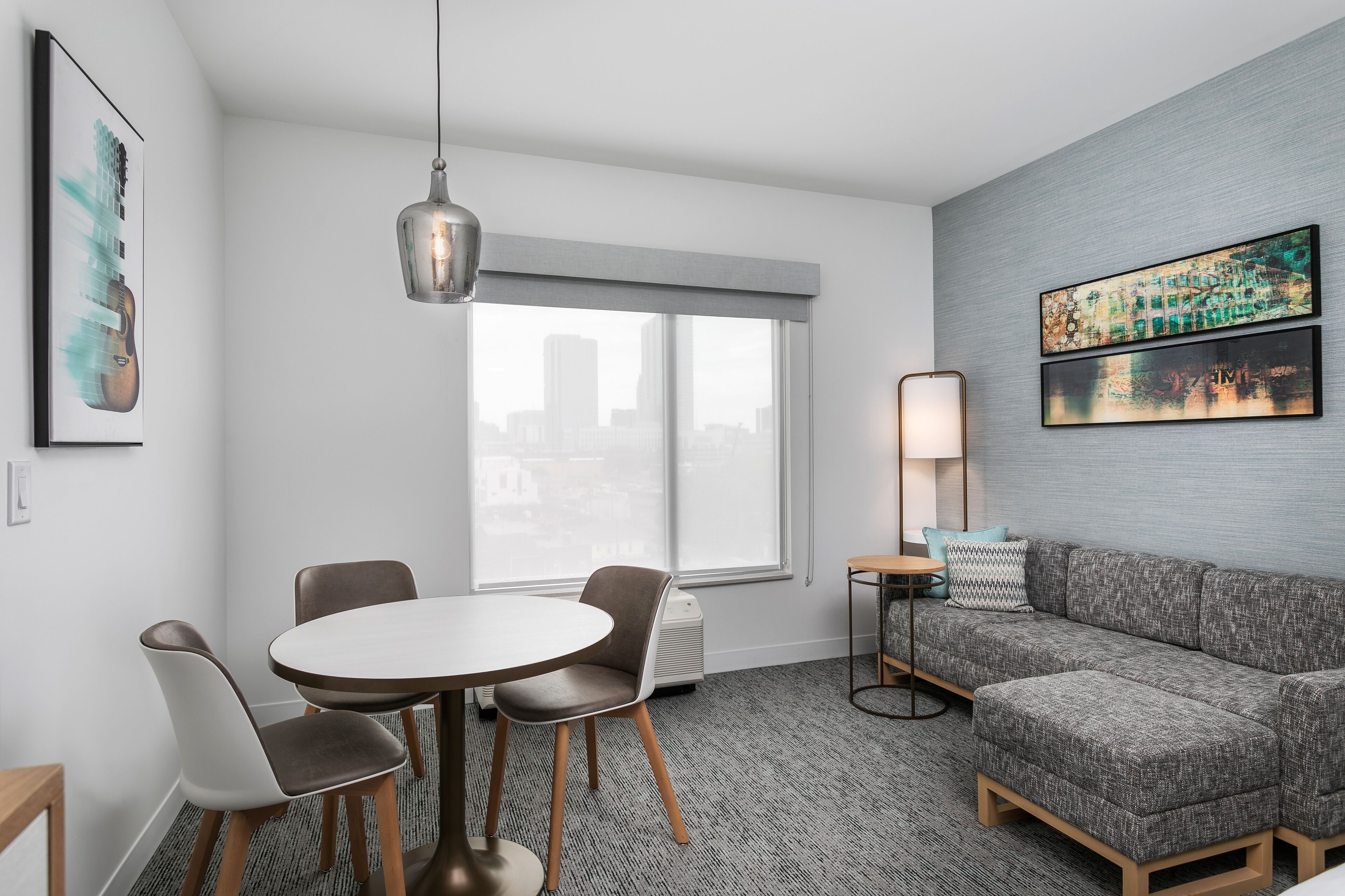 Quarto de hotel moderno com área de refeições, um sofá cinzento e uma grande janela com vista para a cidade.