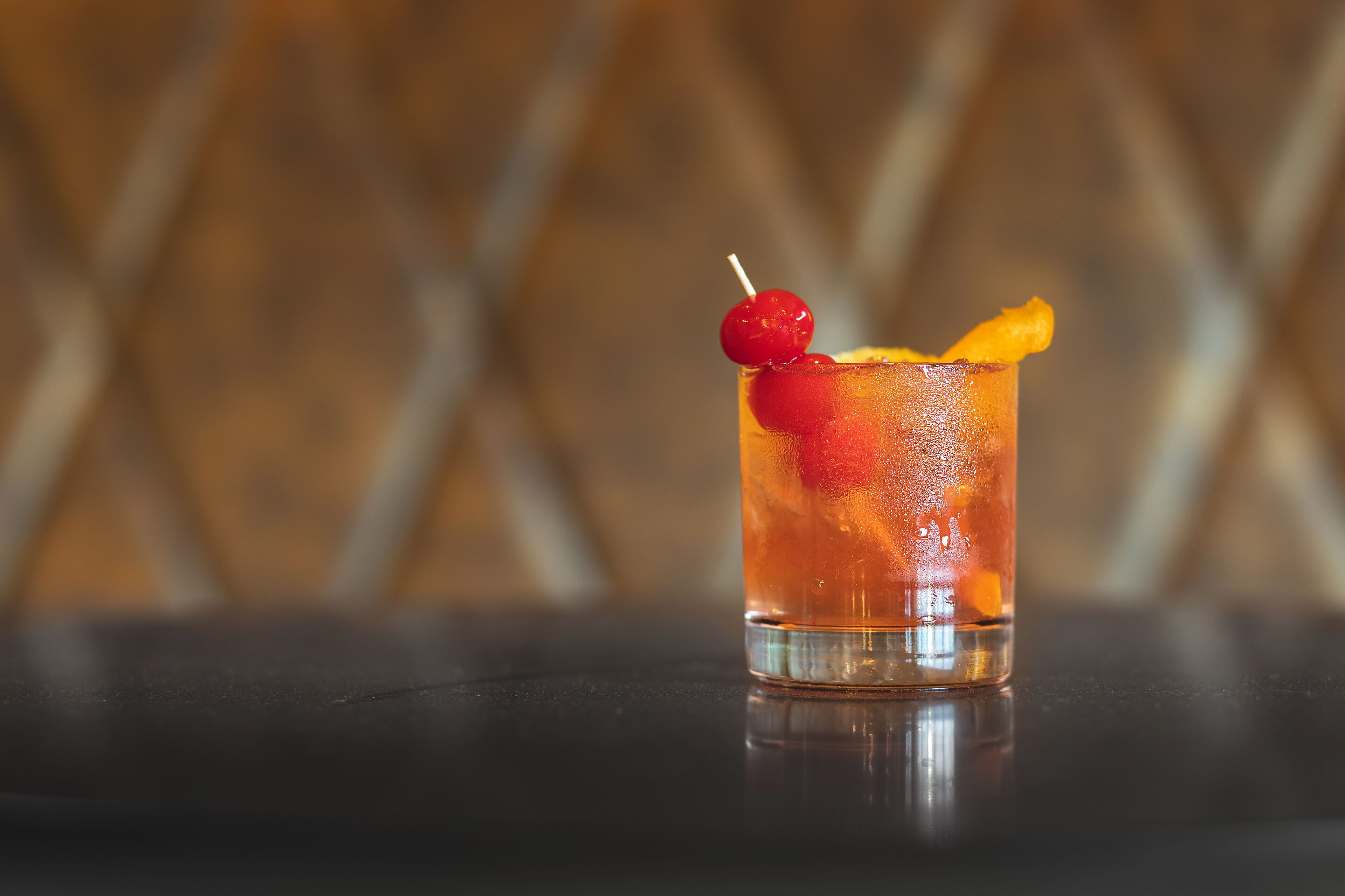 A glass of amber-colored beverage with a cherry and orange slice garnish on a reflective surface.