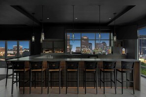 Zona de bar moderna con una barra grande y taburetes, ventanas amplias con vista a la ciudad y lámparas colgantes.