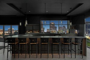 Uma área de bar moderna com uma ilha grande e banquetas, amplas janelas com vista para a cidade e luminárias pendentes.