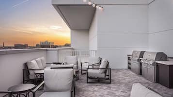 A rooftop patio with outdoor seating, a grill, and a view of the city skyline at sunset.