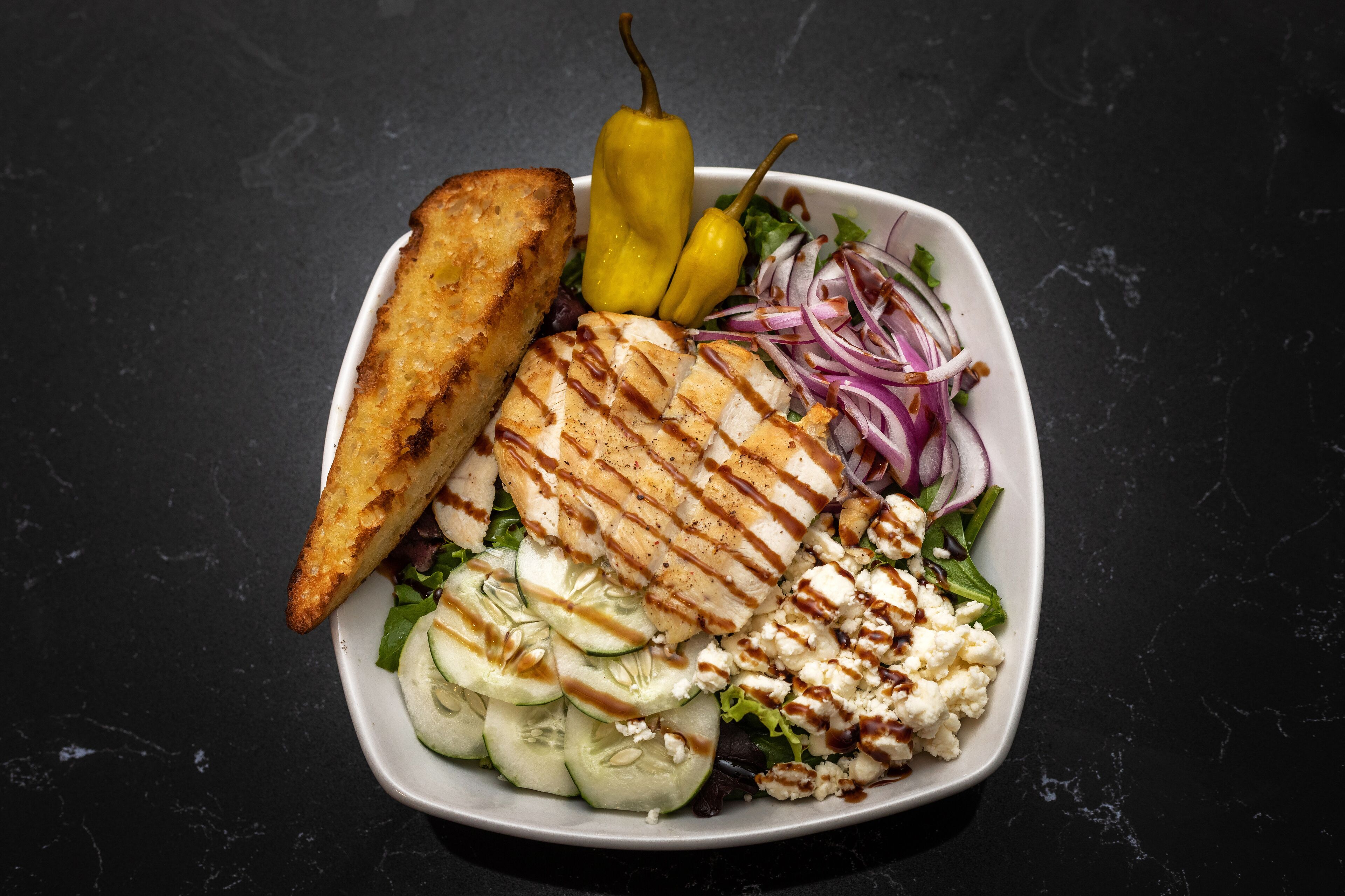 A plate with grilled chicken, feta cheese, cucumber slices, red onion, and a yellow pepper, accompanied by a toasted bread slice.