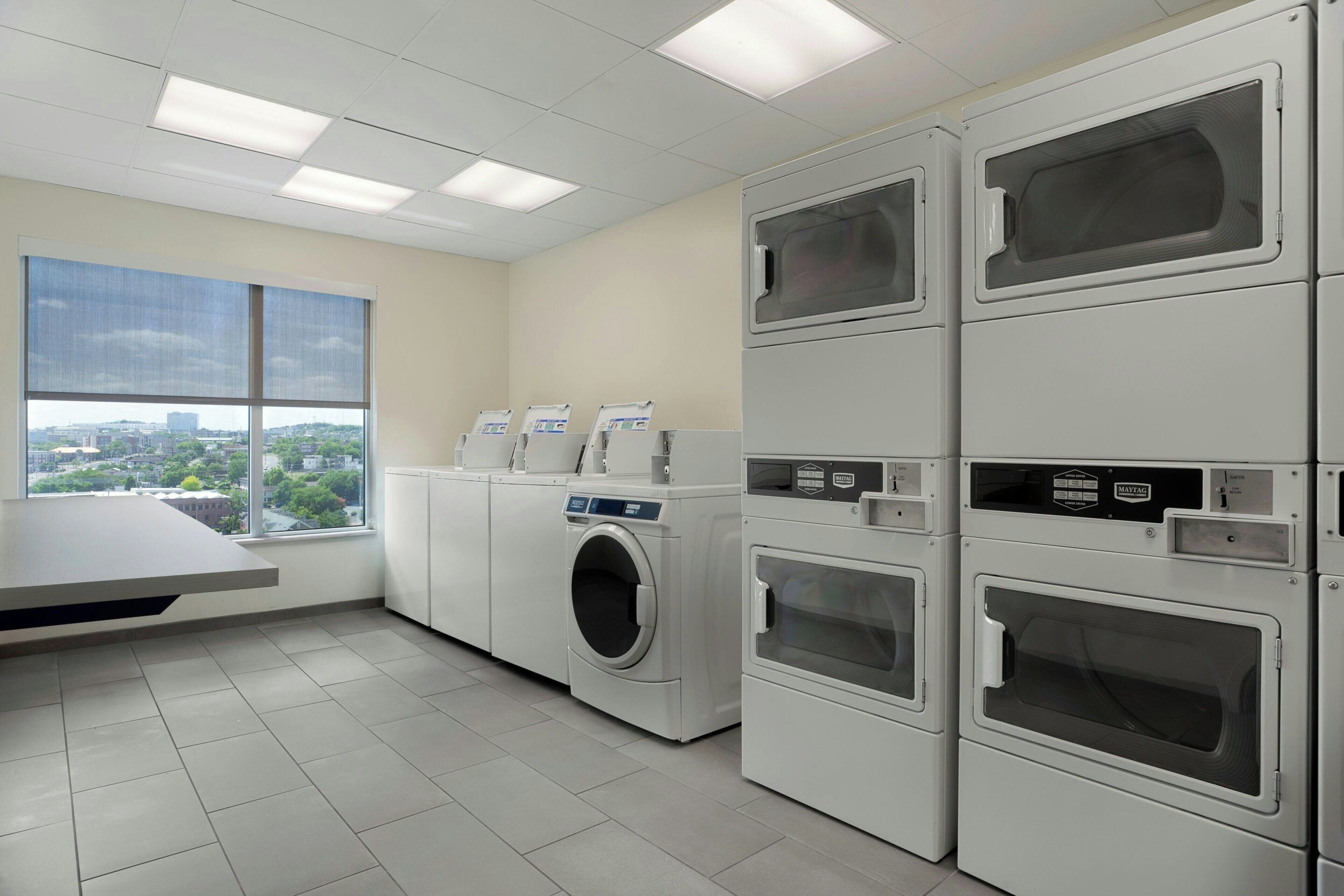 A laundry room with multiple washing machines, a drying rack, and a large window with a city view.