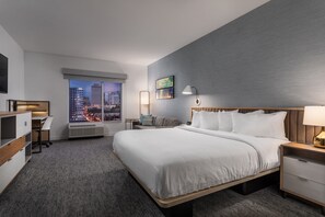 Una habitación de hotel moderna con una cama grande, un escritorio, una silla, una mesita de noche y una ventana con vista a la ciudad.