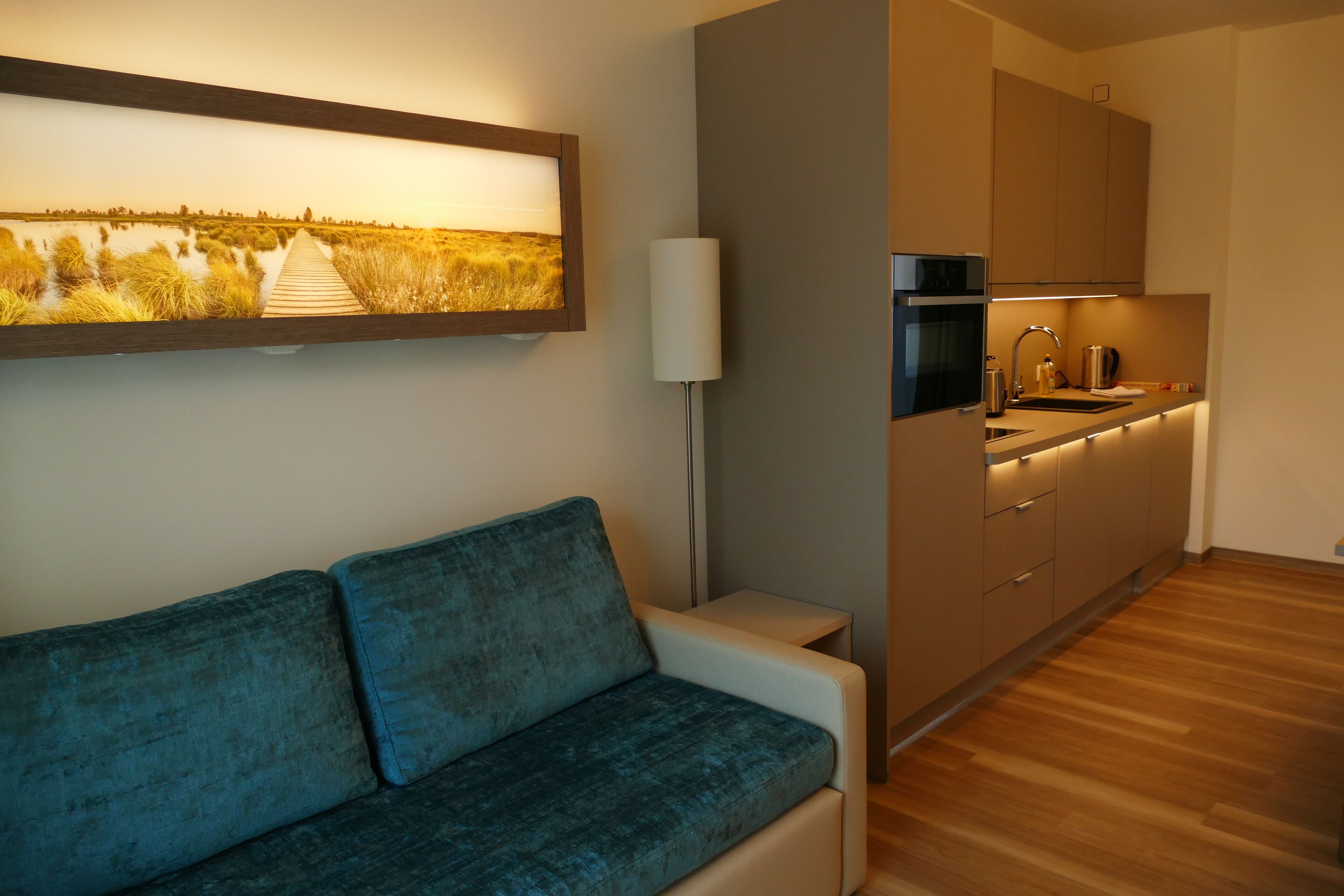 Ein modernes Wohnzimmer mit einem blauen Sofa, Holzboden und einer Küchenzeile mit Holzschränken und einem eingebauten Backofen.