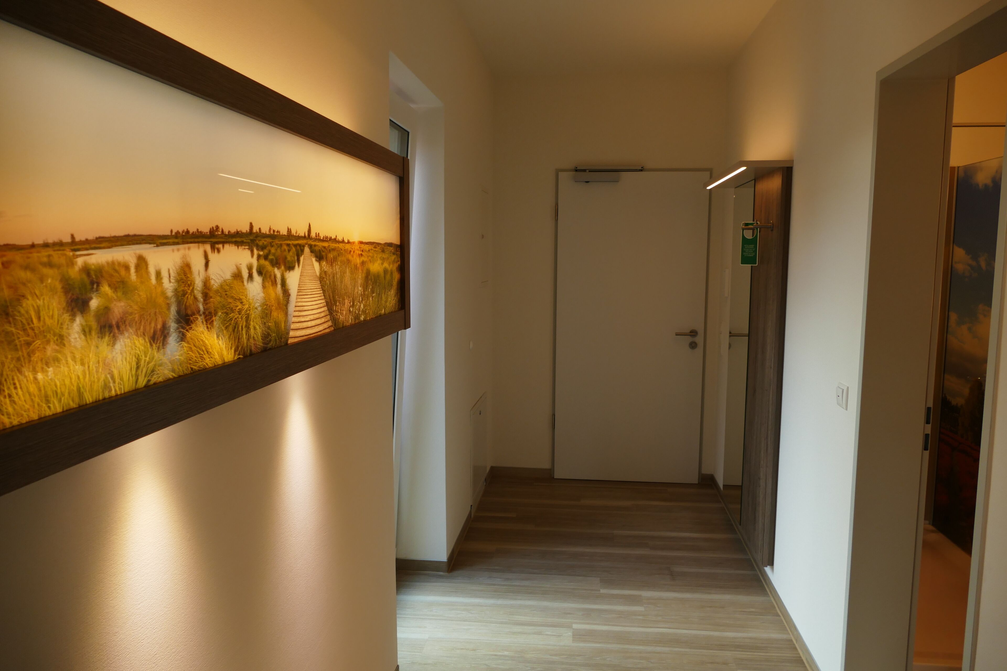 A hallway with a wooden floor, a framed picture of a landscape, and a door at the end.
