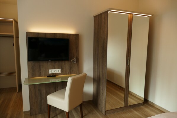 Een moderne kamer met een flatscreen televisie bevestigd aan een houten paneel, een glazen bureau met een stoel en een garderobe met spiegel.