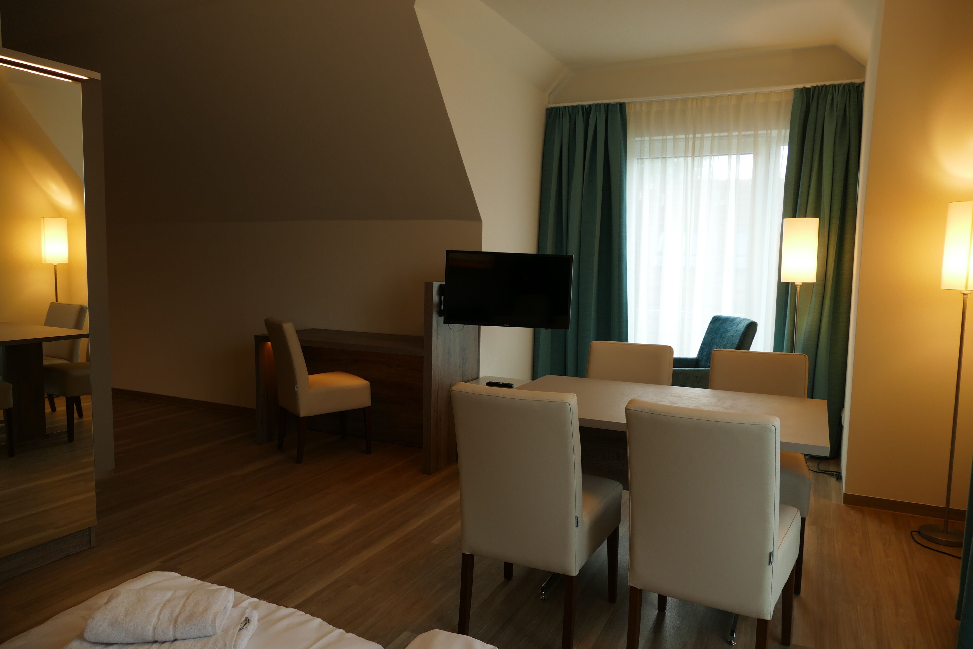 A hotel room with a bed, a desk with a flat-screen TV, a dining area with chairs, and a large window with curtains.