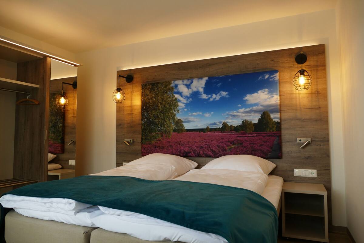 A bedroom with a large bed, a nightstand, a wooden headboard with a painting, and a wardrobe.