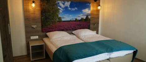 Um quarto com uma cama grande, uma mesinha de cabeceira, uma luminária na parede e uma pintura de uma paisagem.