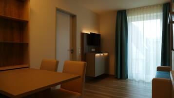 A modern hotel room with a wooden dining table, two chairs, a TV on a stand, a small kitchenette, and a large window with curtains.