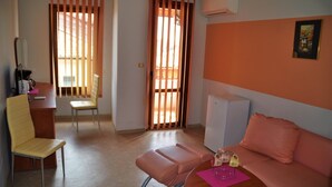 Une pièce comprenant un canapé, une table avec un set de table rose, une chaise, un bureau avec un ordinateur et une fenêtre avec des stores verticaux.
