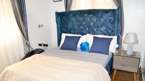 Een net opgemaakt bed met een witte deken en blauwe kussens, een grijze nachtkastje met een lamp, en een blauw gestoffeerd hoofdbord.