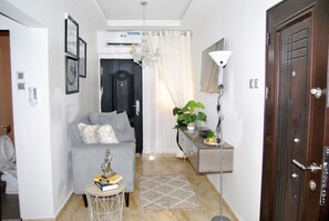 Una sala de estar moderna con un sofá gris, una mesita auxiliar, una araña, una planta en maceta y una puerta de madera.