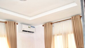 Una camera d'albergo con un letto, tende beige, un condizionatore d'aria e una finestra con tende leggere.