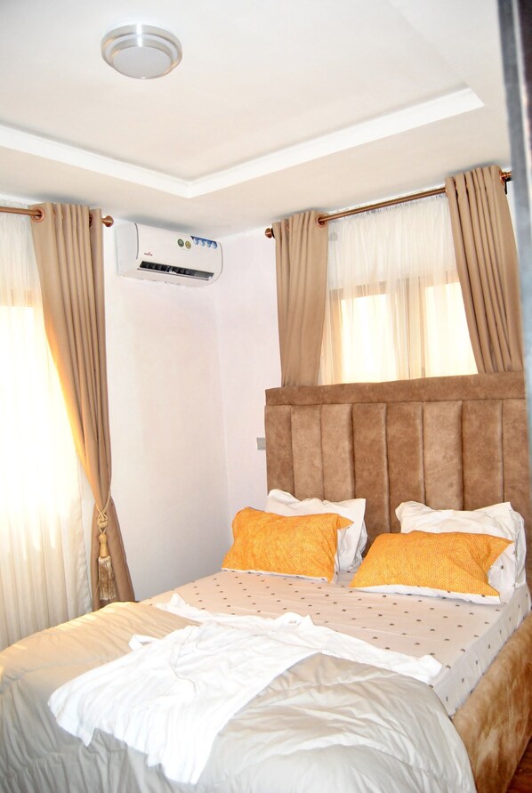 A hotel room with a bed, beige curtains, an air conditioning unit, and a window with sheer curtains.