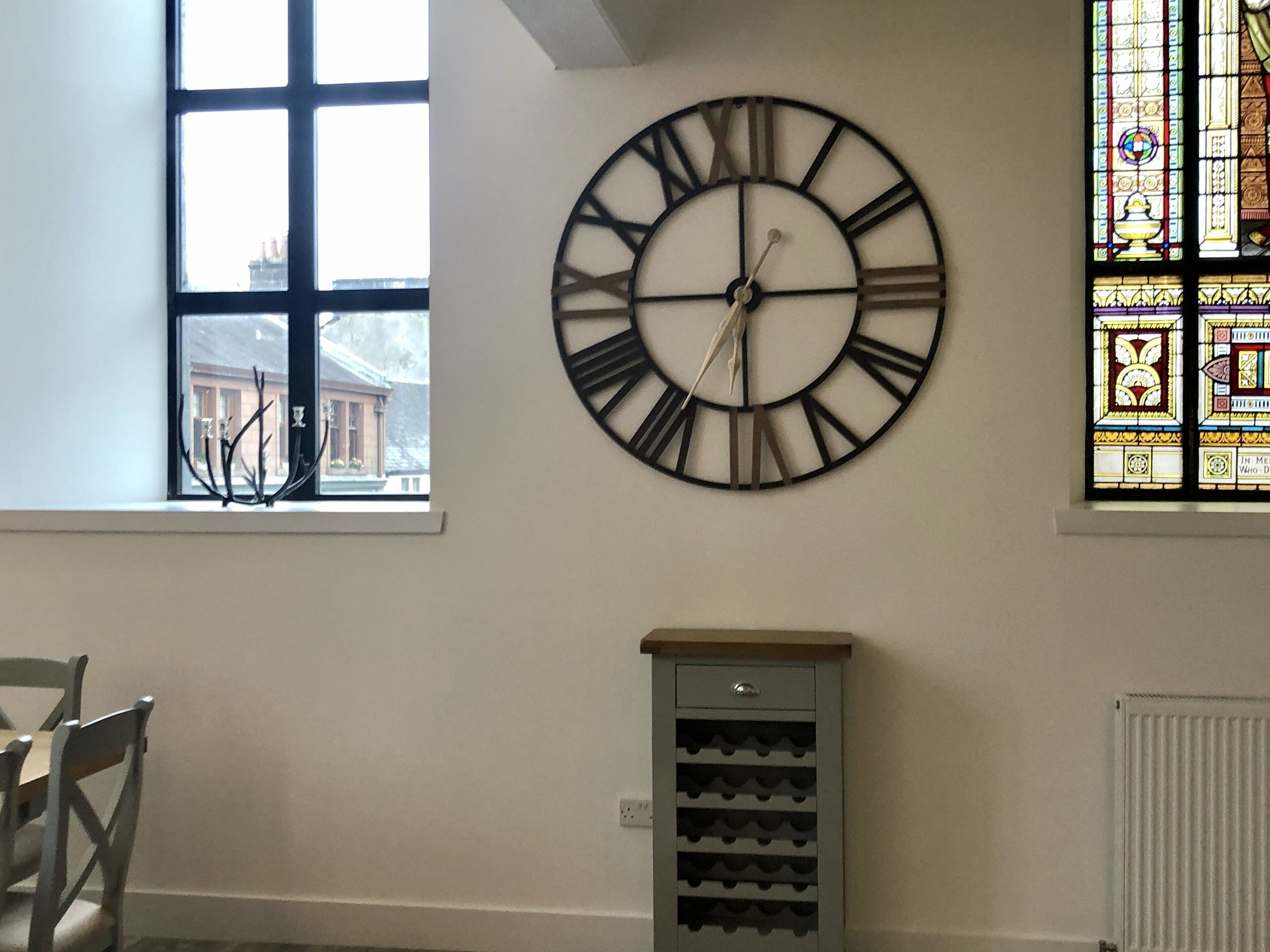 Una habitación con un reloj de pared grande, una ventana con vistas a edificios y una estantería para vinos.