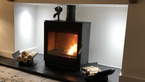 A wood-burning stove with logs and a fire burning inside, placed on a black mantelpiece in a room with a patterned rug. - East Church House (Strathaven)