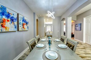 Un espace repas avec une longue table dressée pour six personnes, agrémenté d’un lustre moderne, d’œuvres d’art murales abstraites et offrant une vue sur la cuisine.
