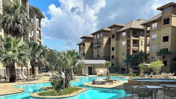 A swimming pool with lounge chairs and a small building, surrounded by palm trees and a multi-story building.