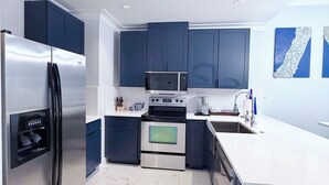 A modern kitchen with stainless steel appliances, dark blue cabinets, a white countertop, and a tiled floor.