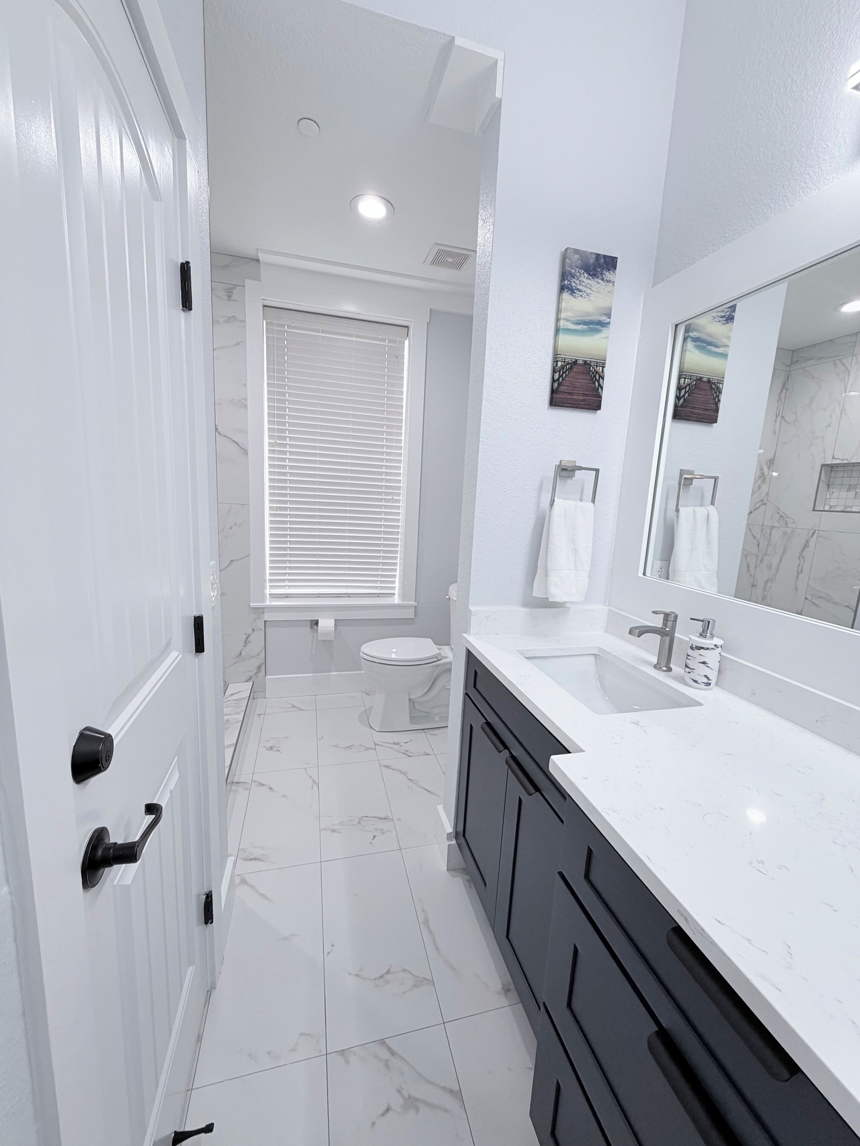 A modern bathroom with a white door, a toilet, a sink with a mirror, and a window with blinds.