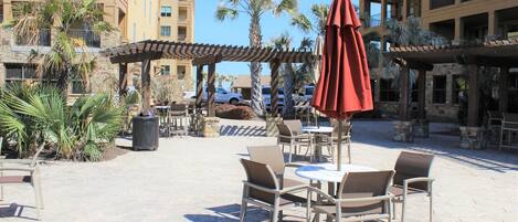 An outdoor patio area with tables and chairs, a red umbrella, and palm trees.