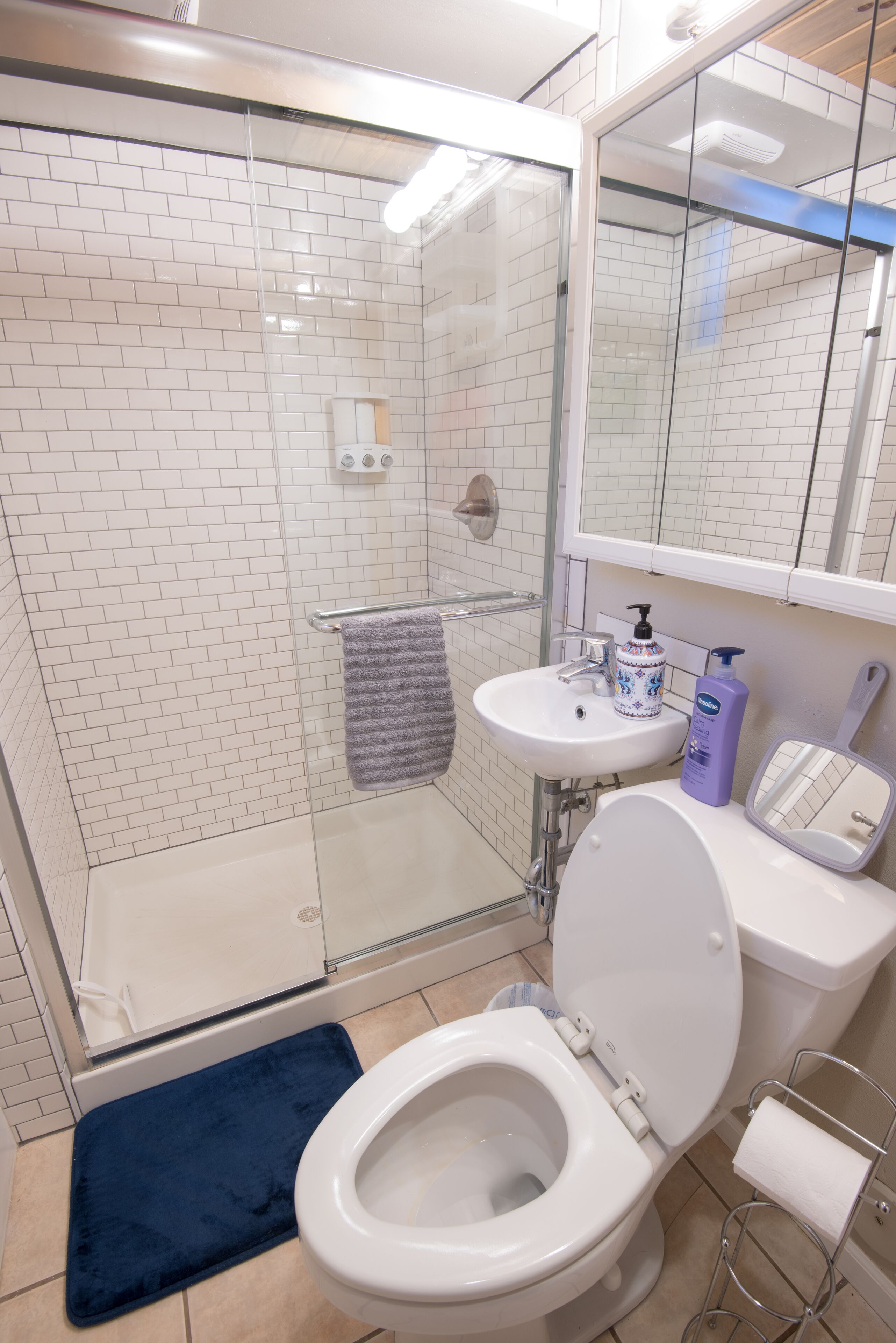 A bathroom with a glass shower enclosure, a white sink with a mirror above it, and a toilet with a blue bath mat in front.