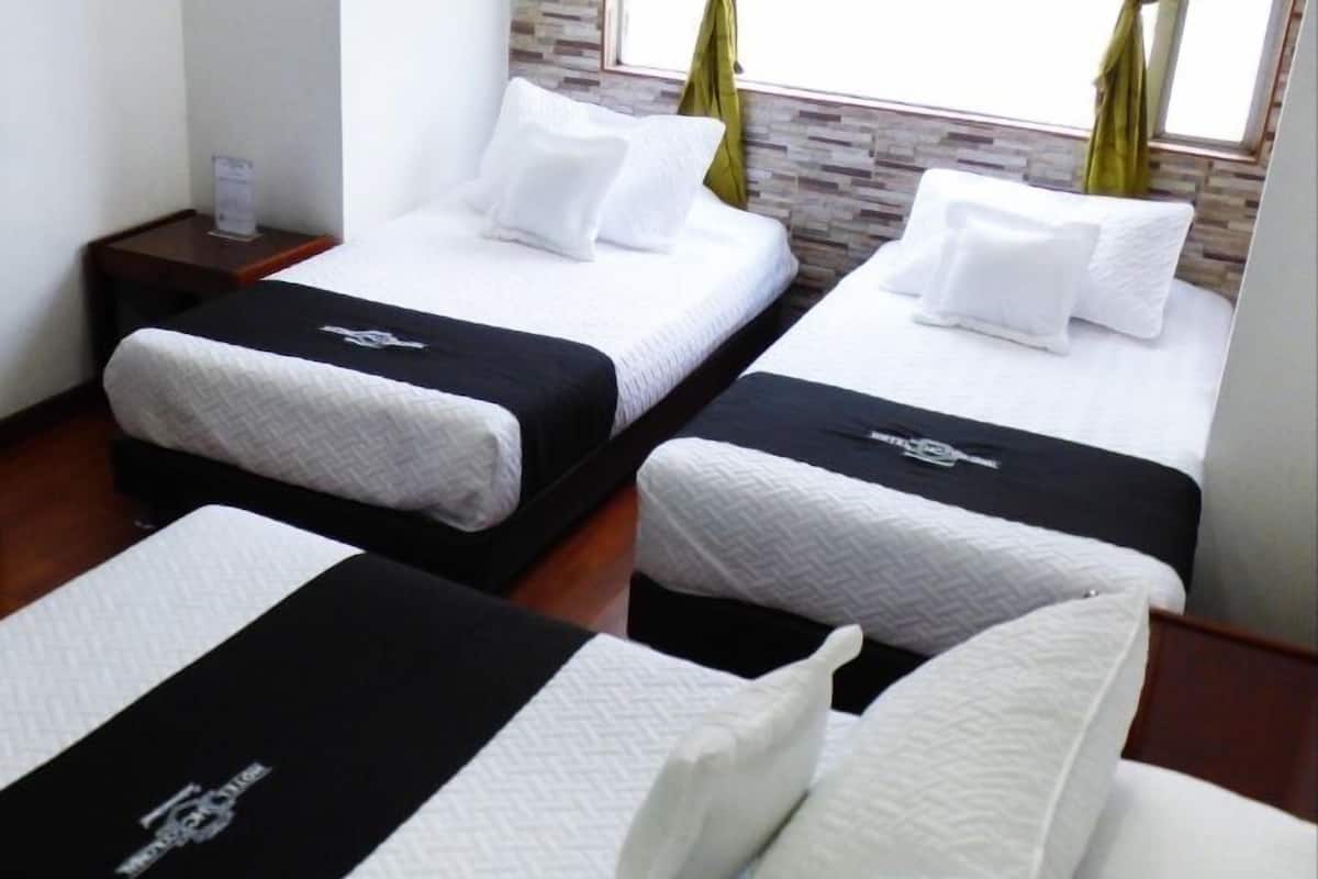 Ein Hotelzimmer mit zwei Betten, jeweils mit weißer Bettwäsche und einem schwarzen Kopfteil. Das Zimmer hat einen Holzboden, eine Wand in Backsteinoptik und grüne Vorhänge an den Fenstern.