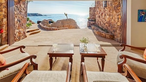 Una habitación con pared de piedra y vistas al mar. Hay dos mesas de madera con sillas, un florero con flores y una planta en maceta.