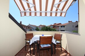 A terrace with a table and chairs, a striped awning, and a view of buildings in the background. - Apartments Darko (Rovinj)