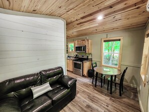 A cozy living room with a leather sofa, a dining table with chairs, a kitchen with wooden cabinets, and a large window with a view of greenery.