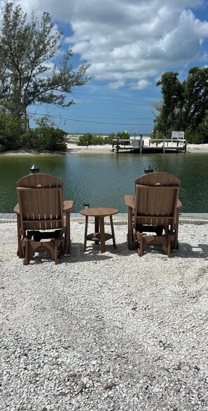 Two wooden Adirondack chairs and a small table on a gravel surface by a body of water, with a dock and a boat in the background.