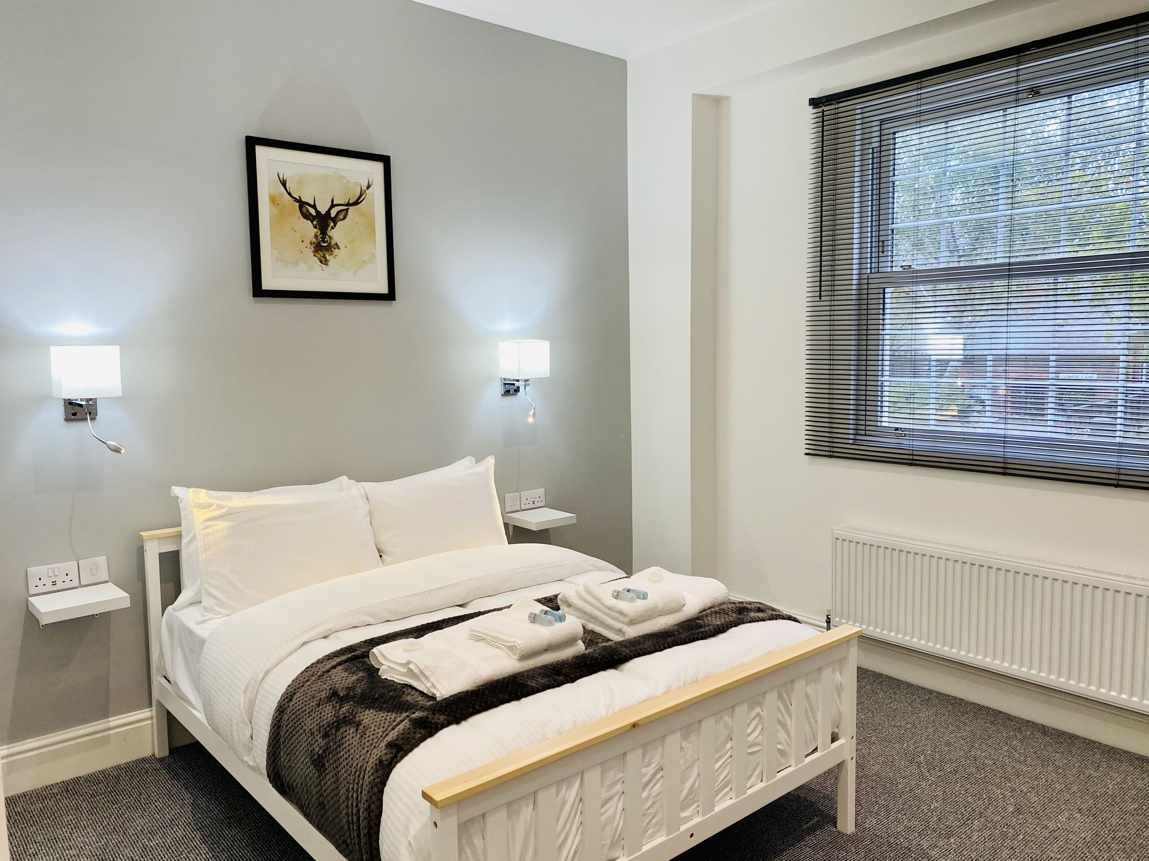 A neatly made bed with white linens, a dark patterned throw, and a white headboard. There are two bedside tables with lamps, a framed deer head artwork on the wall, and a window with blinds.