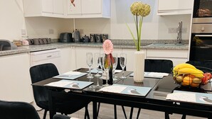 A modern kitchen with white cabinets, a stainless steel range hood, and a dining area with black chairs and a glass table. - Higher Lane House (Plymouth)